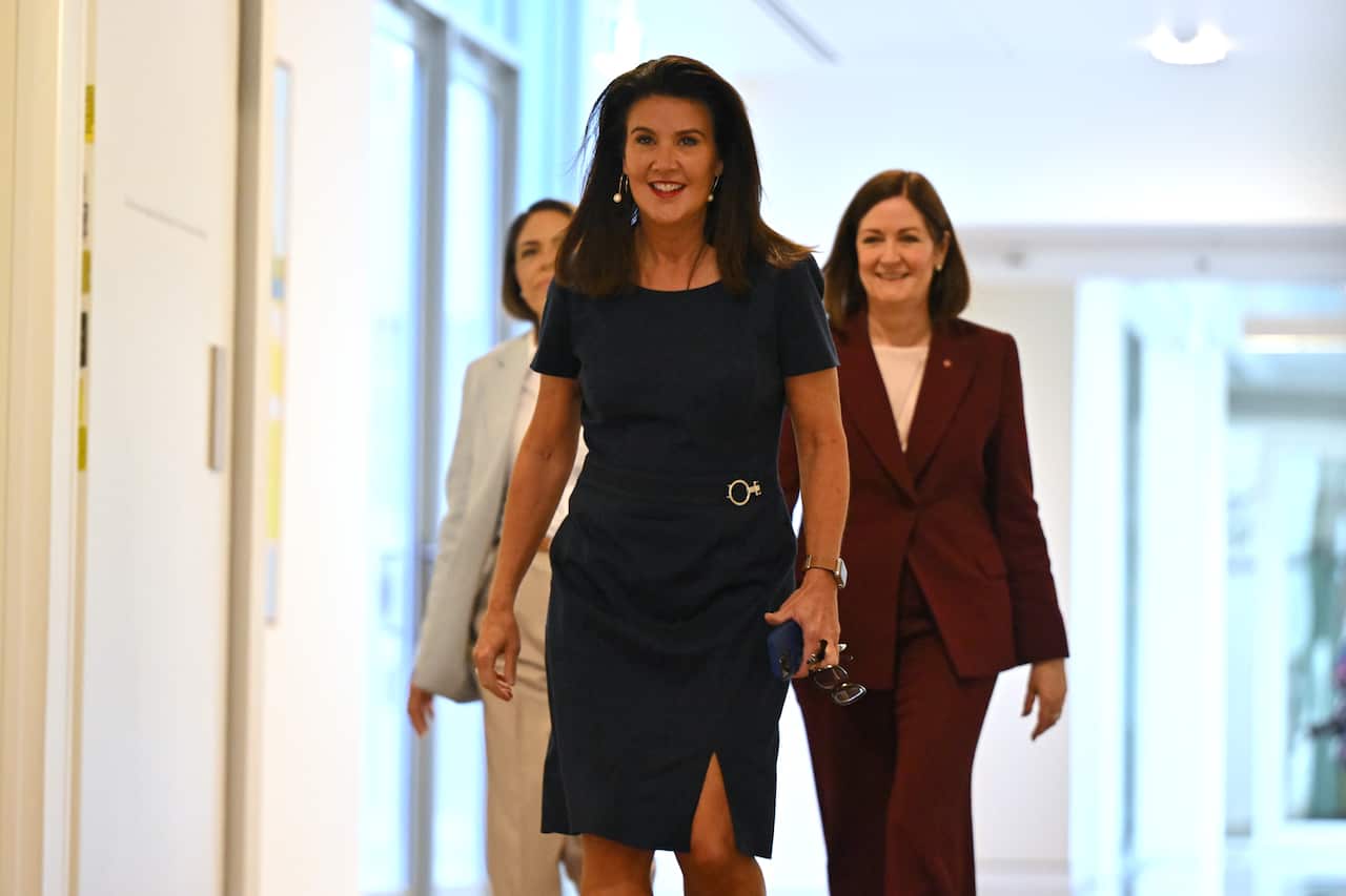 A white woman in a dark dress walking down a corridor, two other women are behind her 