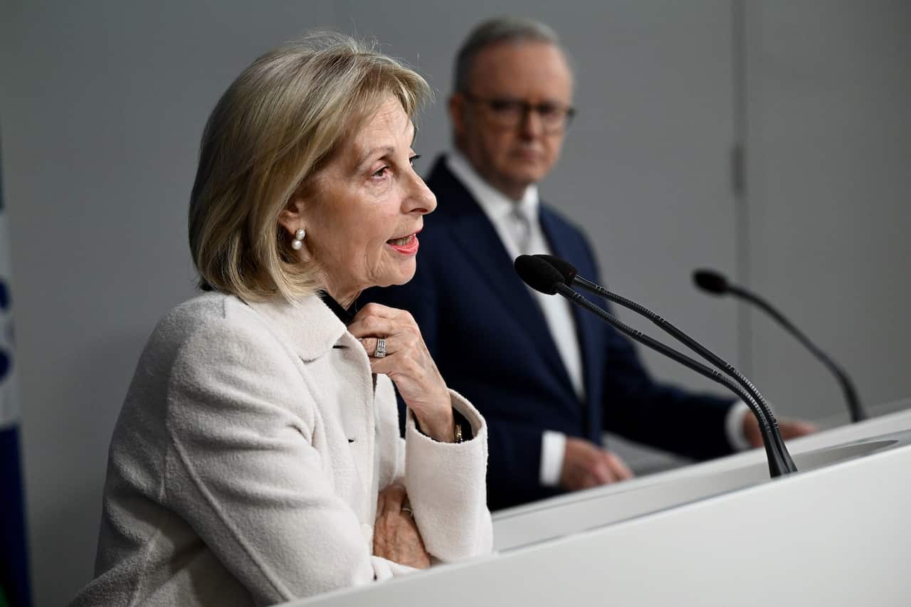 A woman in a white blazer speaks into a microphone. A man in a dark suit, standing next to her, looks at her.
