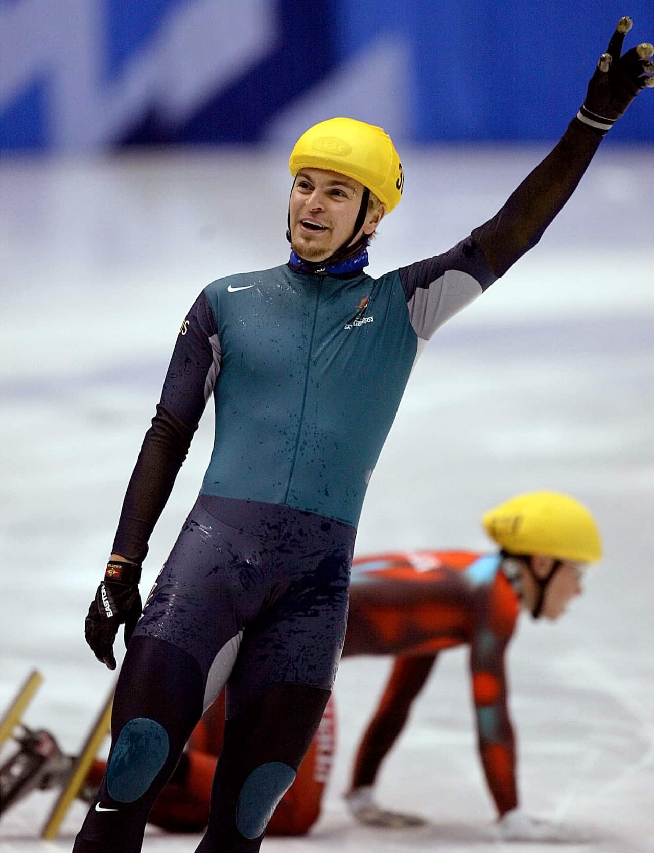 An ice skater wearing a tight bodysuit that is green in the body and black on the legs and a yellow helmet holds up his left arm above his head.