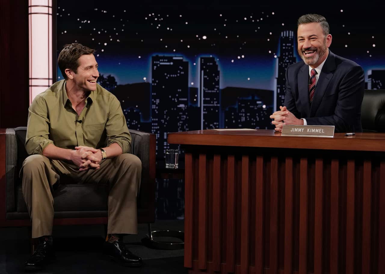 Two white men on a laste night TV show set. One is sitting on a chair, the other is seated behind a desk.