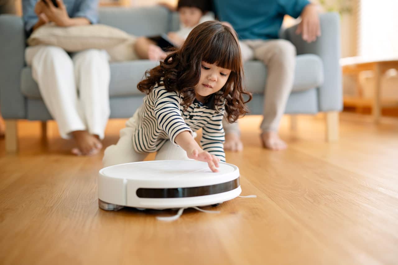 Get Smarter and Safer in Home Automation Technology. A child girl looked excited is switching on a robot vacuum to auto cleaning on a hardwood floor in the living room while her parent is controlling the vacuum device via a mobile app.