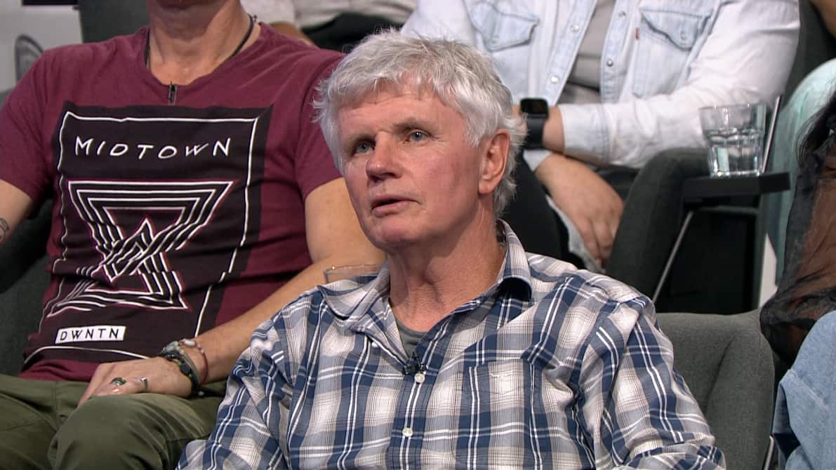 A screenshot of a man in a checked shirt sitting in a TV studio.