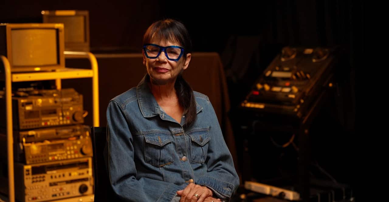 A woman wearing blue glasses and a denim shirt is sitting in a dimly lit studio.