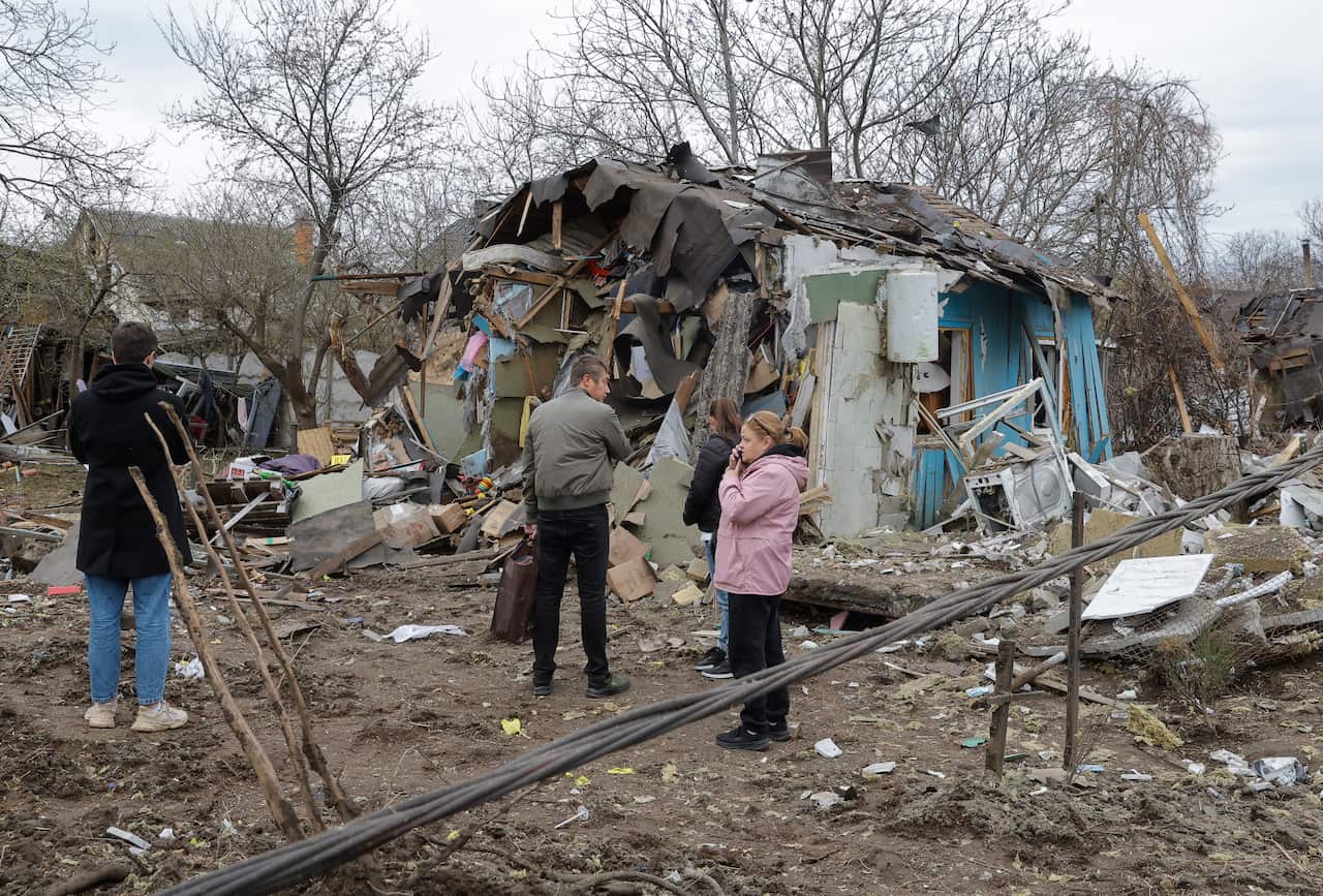 Four people stand amidst the debris of a heavily damaged, bright blue house that has been partially leveled by an explosion or conflict.