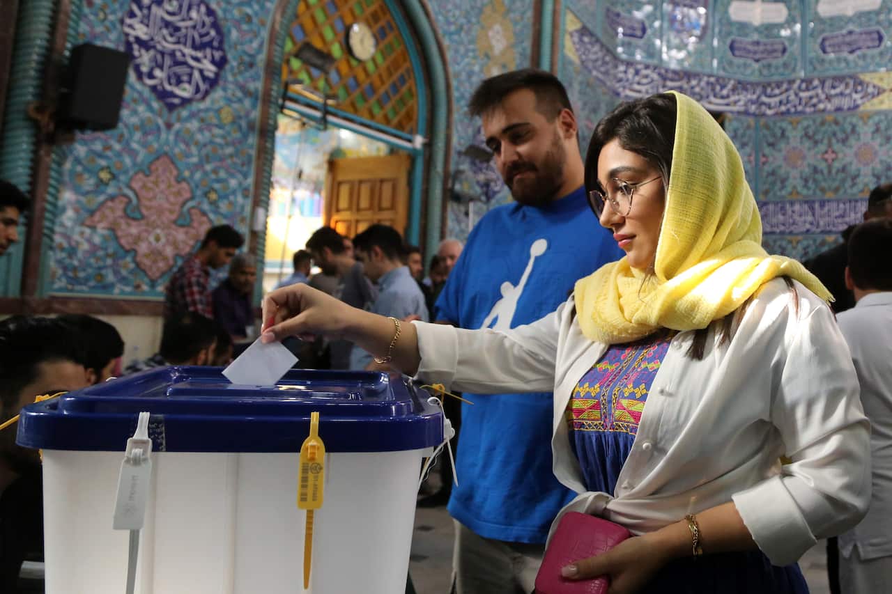 A woman putting a piece of paper into a ballot box at a polling station. A man in a blue shirt is behind her.