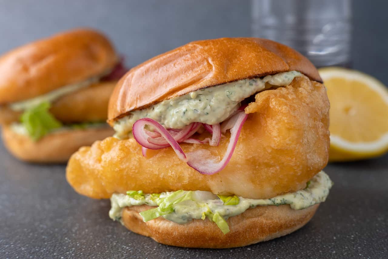 A thick golden battered fish fillet sits in a bud, with a green-speckled sauce and rings of pickled red onion. A second burger can be seen sitting behind.
