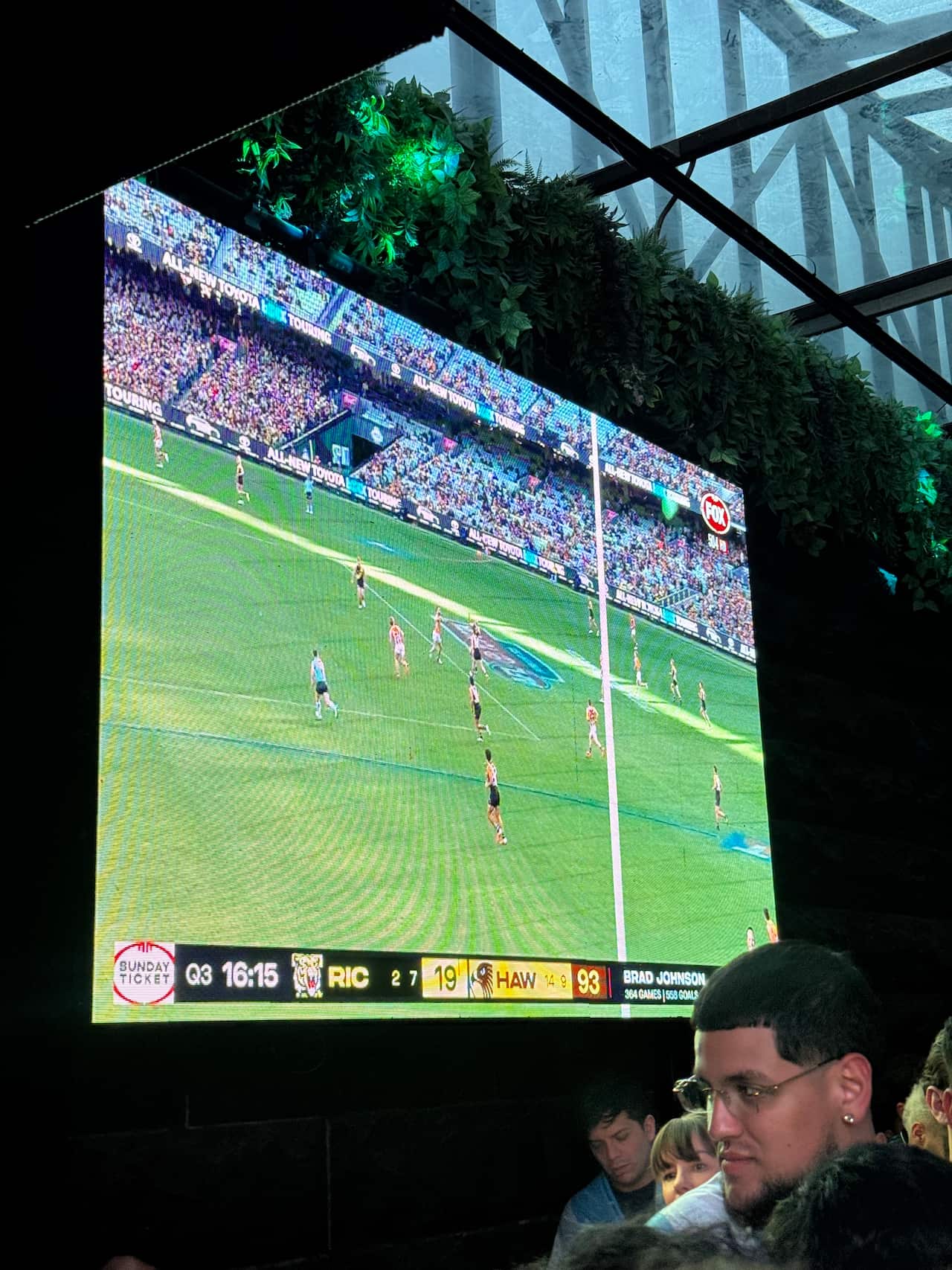 A large screen at a public venue shows an AFL game in progress.