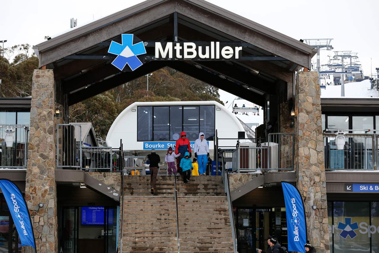 A large stone and wood arch with a sign saying Mt Buller at the top. There is a staircase going through the arch on which five tourists are walking.