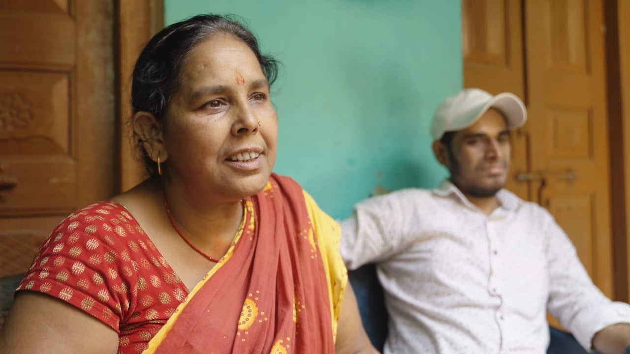 A woman in a red and yellow traditional Indian sari sitting next to a younger man wearing a white shirt and a white baseball cap