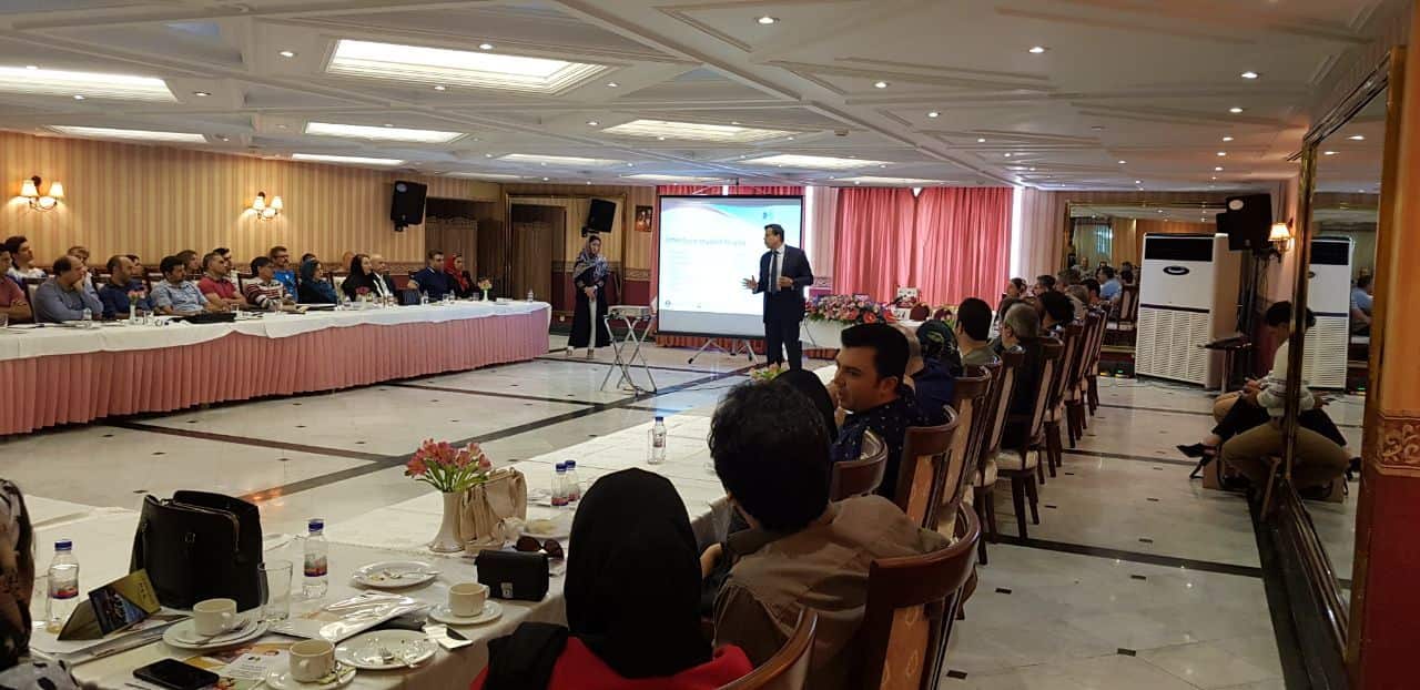 A presenter in a suit stands on a stage next to a large projection screen, addressing an audience seated at long tables. The seminar room has a decorative ceiling, a patterned floor, and pink drapes at the front.