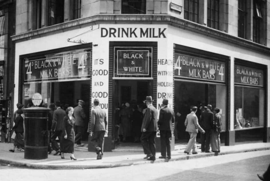 The outside of a milk bar on a street corner, with passing pedesrians.