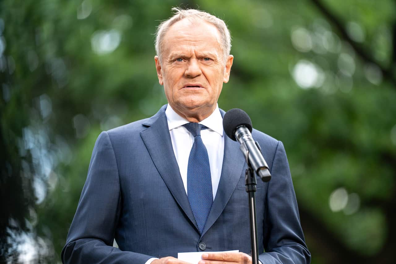 Donald Tusk at a press conference in a park on a hot day, wearing a blue suit and standing in front of a microphone stand.
