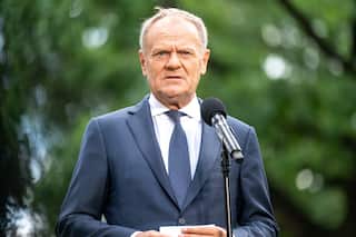 Donald Tusk at a press conference in a park on a hot day, wearing a blue suit and standing in front of a microphone stand.