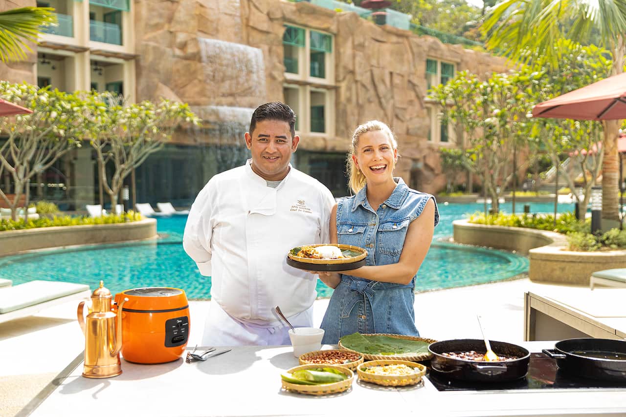 A man in chef's whites and a smiling woman stand in front of a resort pool. A table in front of them holds a cooktop and various ingredients. The woman holds up a platter full of food. 