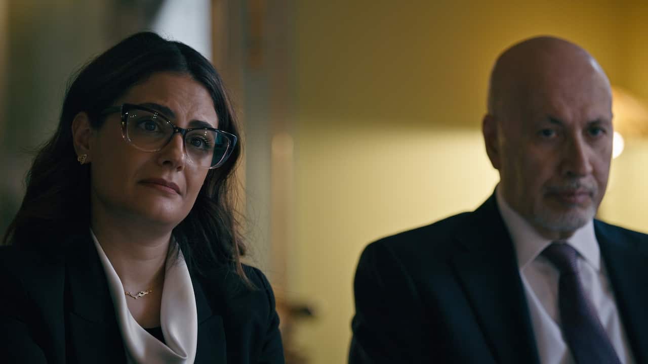 A woman with dark hair and glasses looks on with a concerned expression while seated next to a man in a suit and tie.