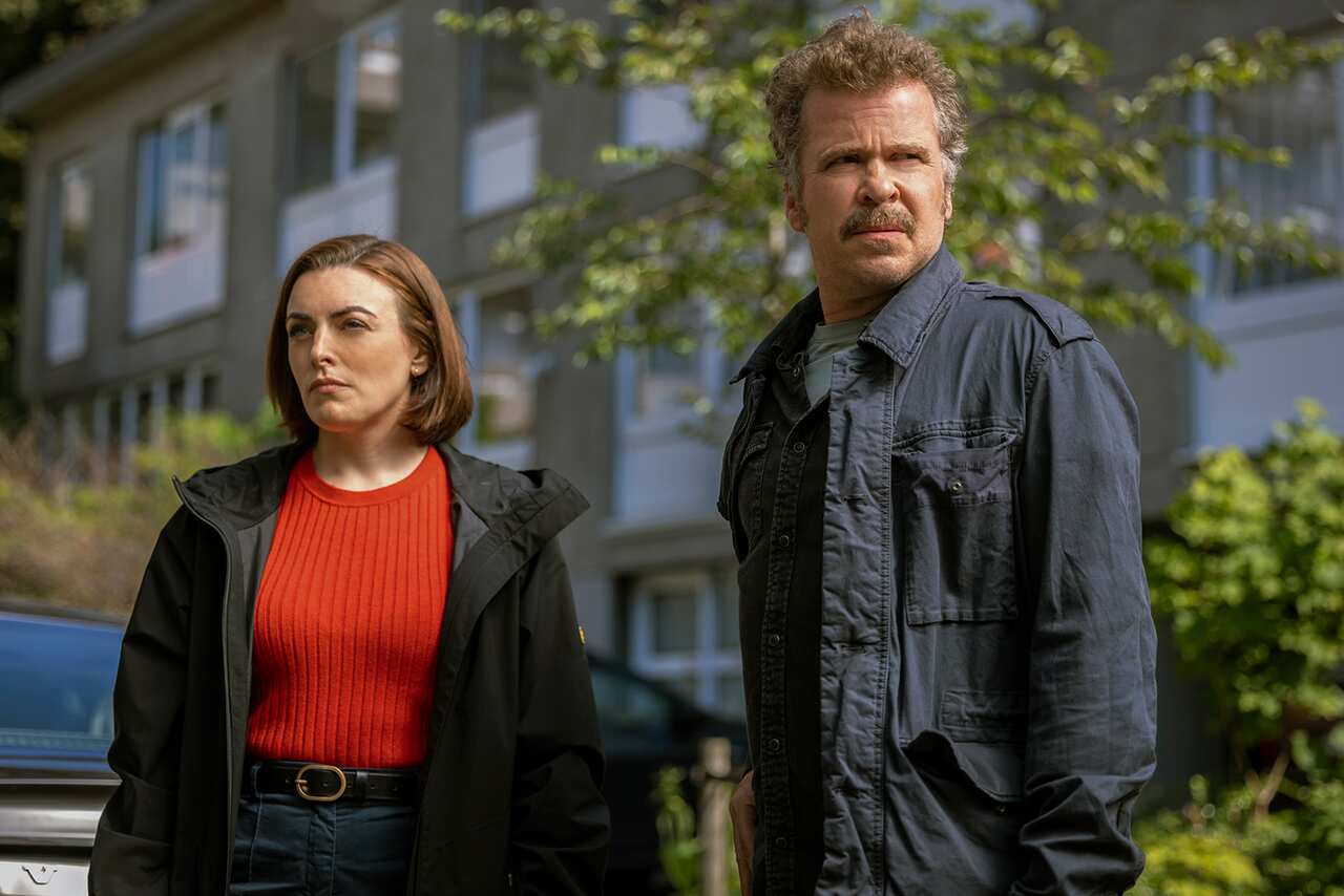 A woman in a red shirt and dark coat and a man in a dark shirt stand in front of a building. They are looking off in different directions, both with serious expressions. 
