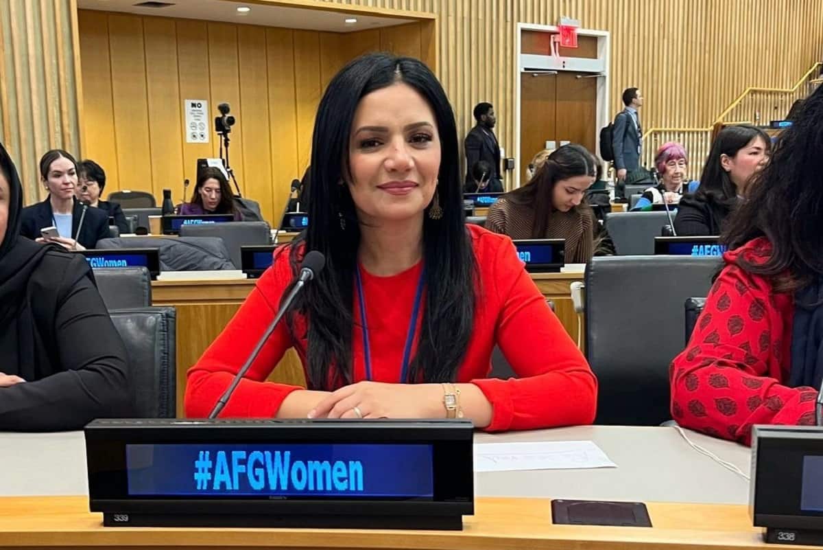 A woman in a red top and with long black hair sits on a long table in front of a microphone and a small black screen panel, with "#AFGWomen" displayed on it.