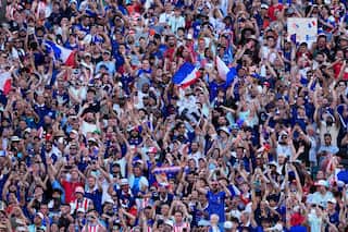A crowd of French World Cup fans wearing red, white and blue raise their arms and flags in excitement.