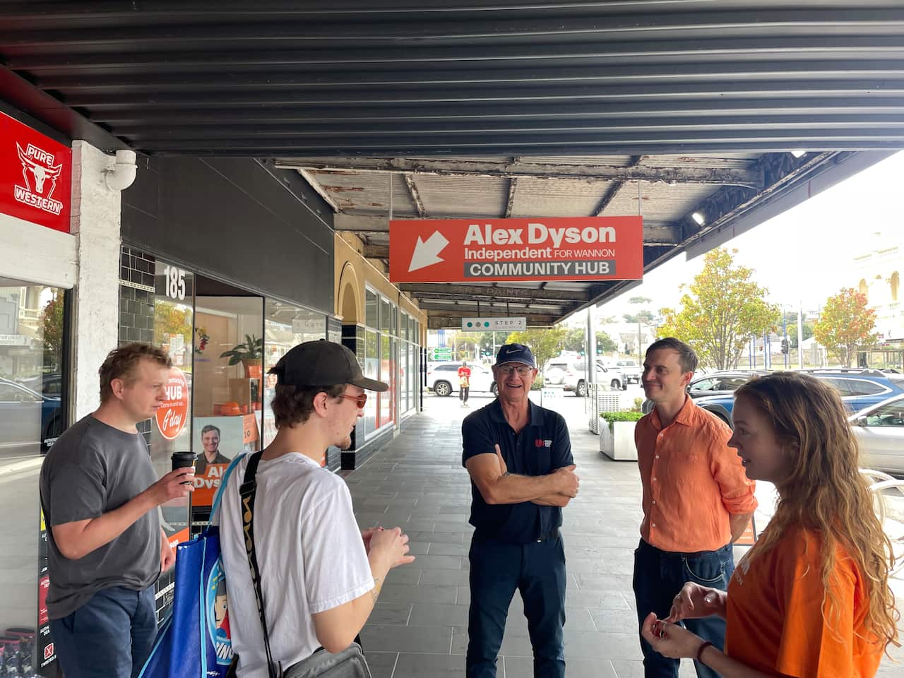 A man in an orange shirt is speaking to a group of people outside shopfronts.