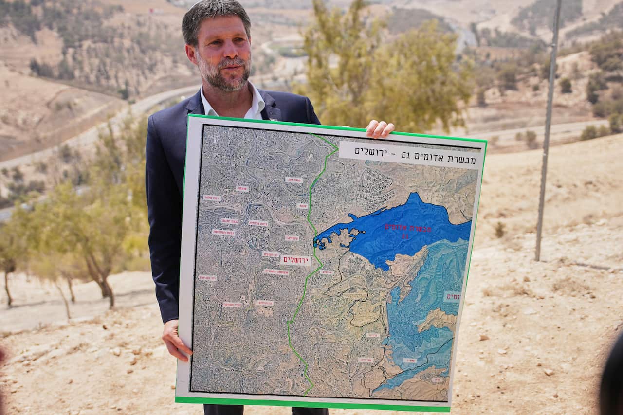 A man in a dark suit holds a large sign with a map, behind him is sand and trees.