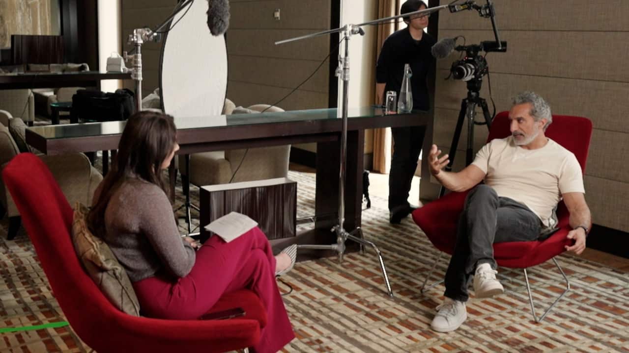 A woman and man sit across from each other in red chairs in a hotel room, with a camera operator and camera beside them.