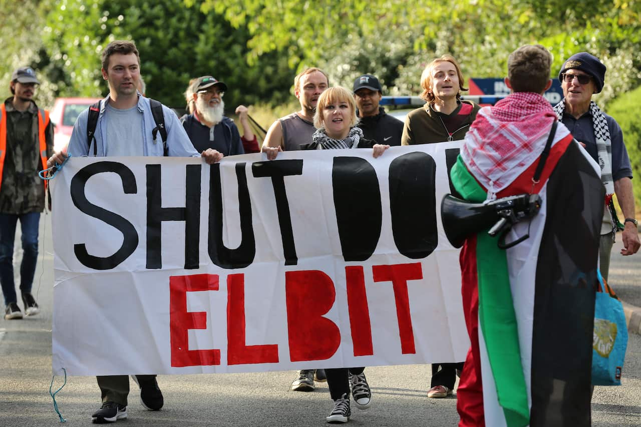 A group of people, one draped in a Palestinian flag and megaphone, hold a banner reading Shut Down Elbit.