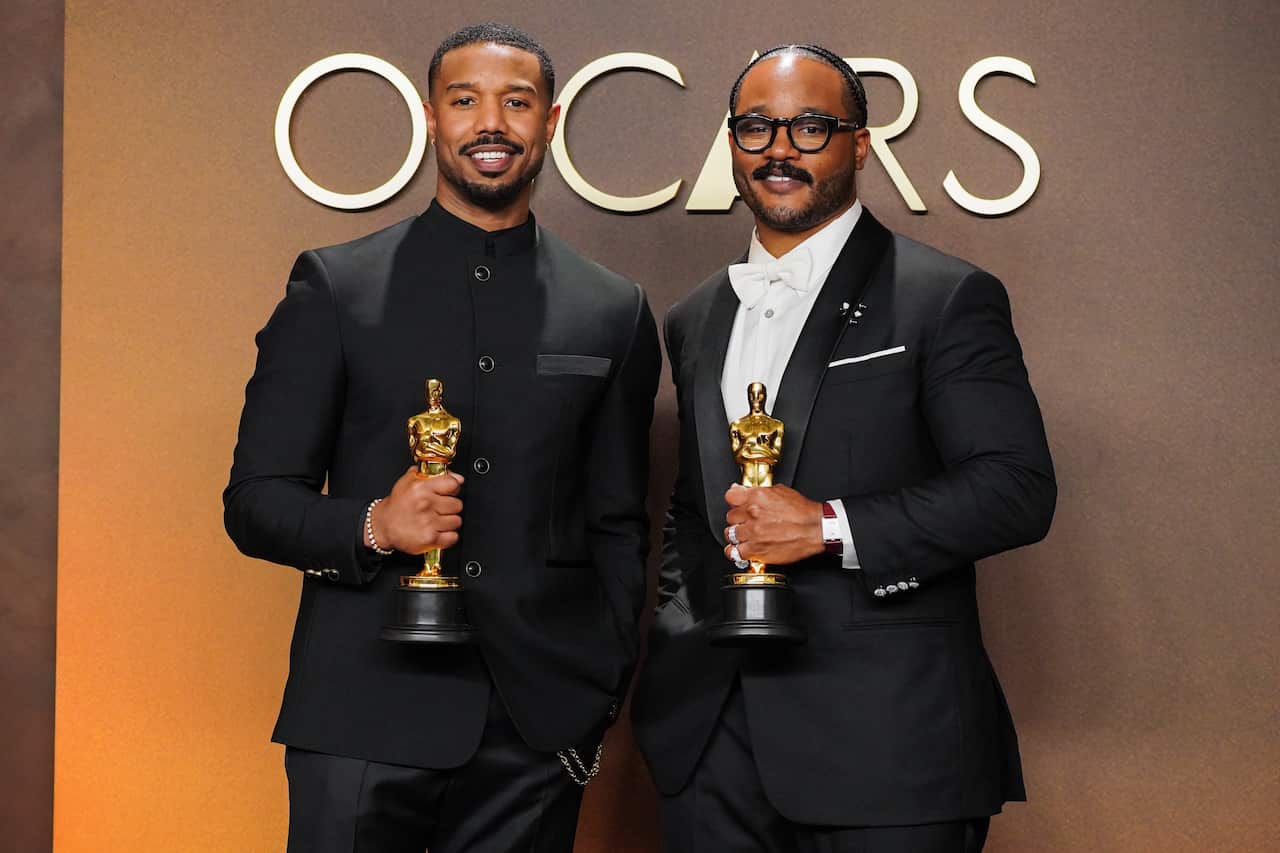 Michael B Jordan and Ryan Coogler, standing next to each other and holding up Oscar awards.