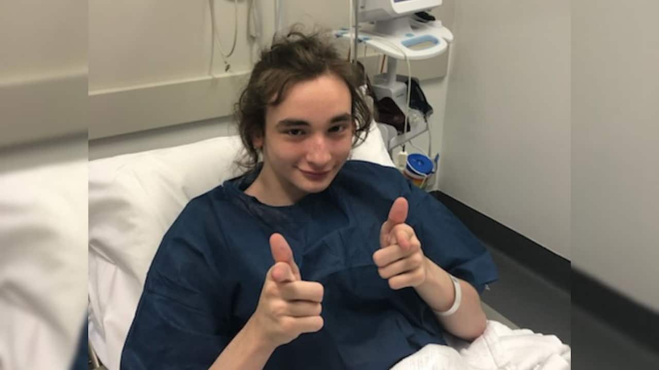 a young man with long brown hair does finger guns to the camera from a hospital bed