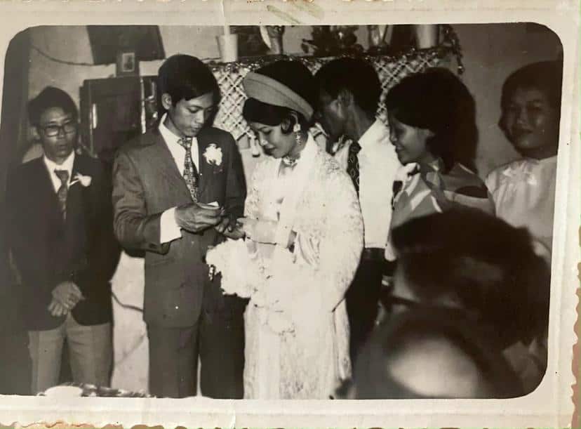 Black and white photo a man in a suit standing next to a woman wearing a white dress and a dark headband. There are people looking on
