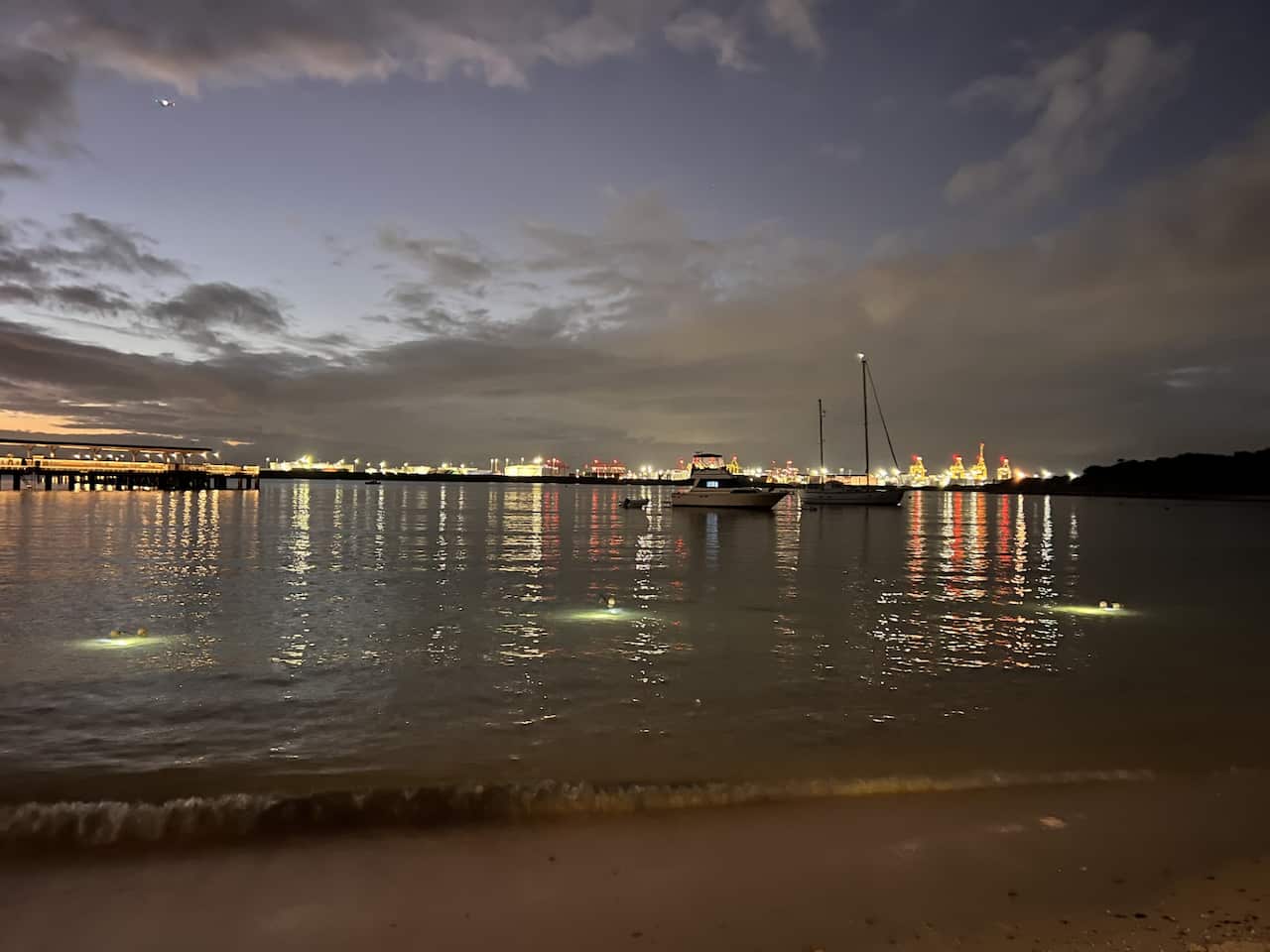 A wide shot of a bay, capturing the expansive sky, the body of water, and urban lights, with some boats visible at a distance.