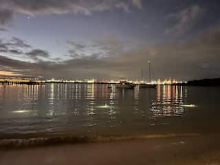 A wide shot of a bay, capturing the expansive sky, the body of water, and urban lights, with some boats visible at a distance.