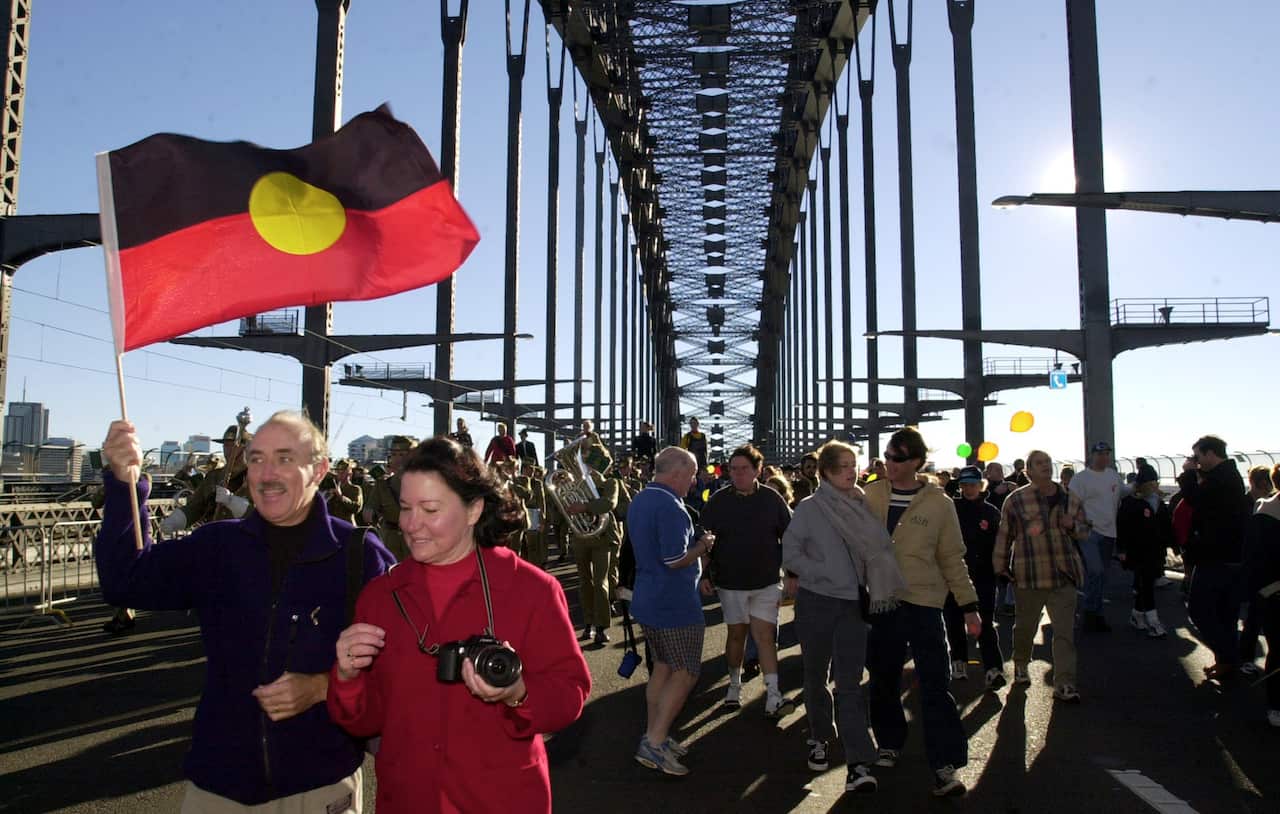 People walking across the Sydney Harbour Bridge with a man holding an Aboriginal flag in front.