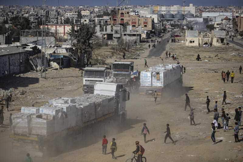 An aerial view of two trucks loaded with white cartons driving on dusty land. People look on