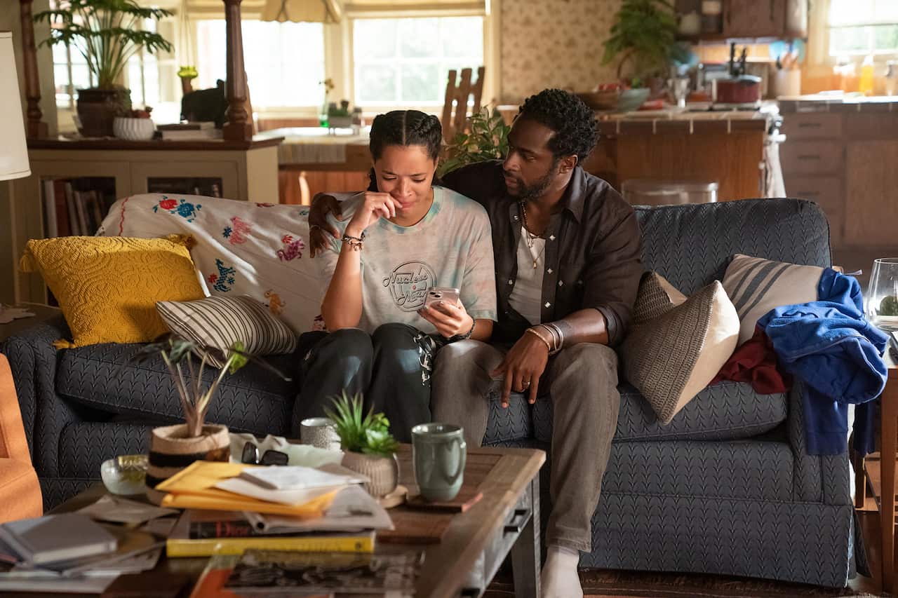 A man and a young woman sit on a couch. She holds a phone and looks upset, he has his arm around her, comfortingly.