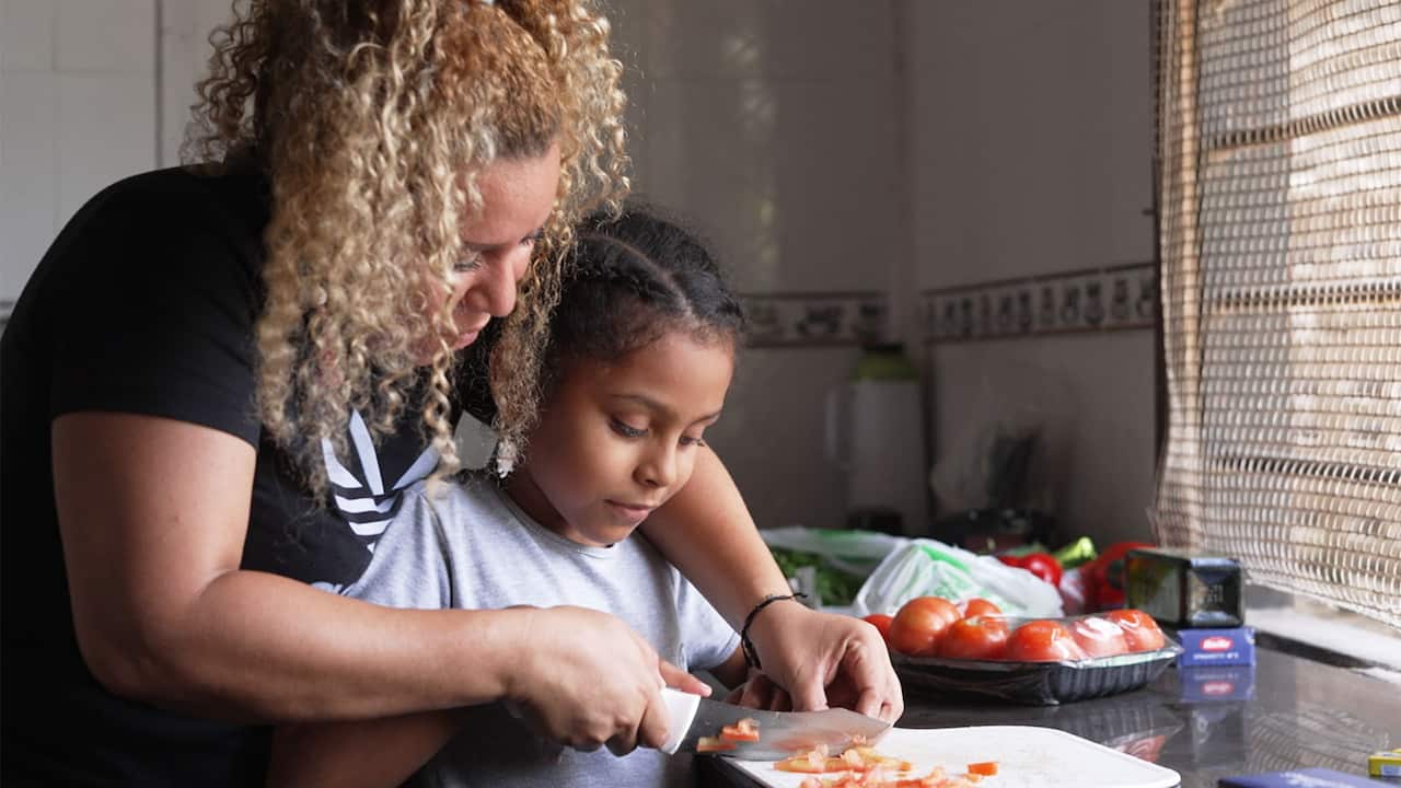 A woman in black t-shirt and high ponytail embracing a young girl from behind as they slice tomatoes on a chopping board