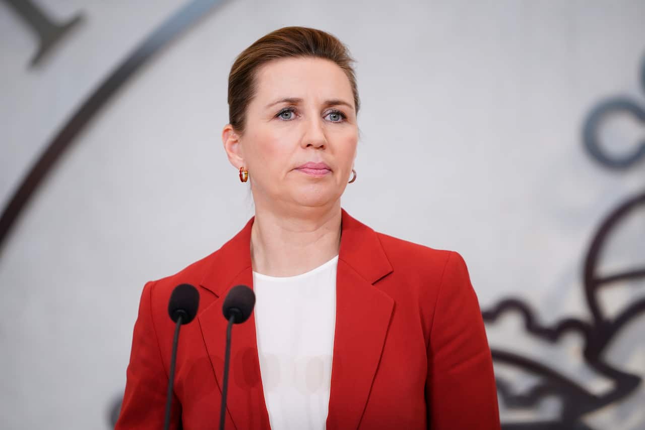 A woman in a white top and a red blazer stands in front of two microphones.