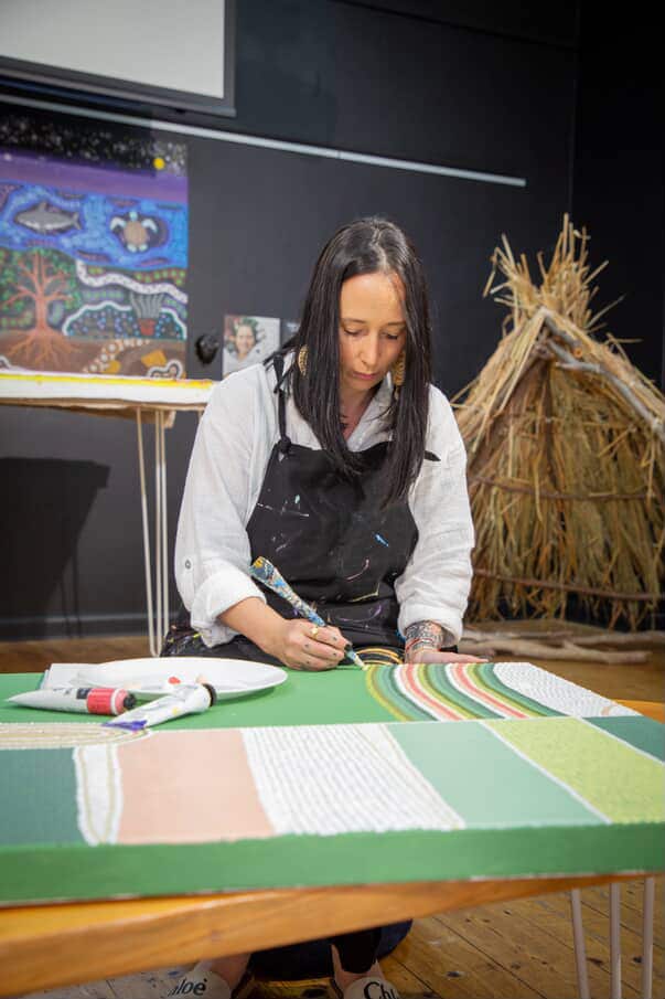 A woman with dark hair wearing a white shirt and dark apron is behind a table painting a green canvas