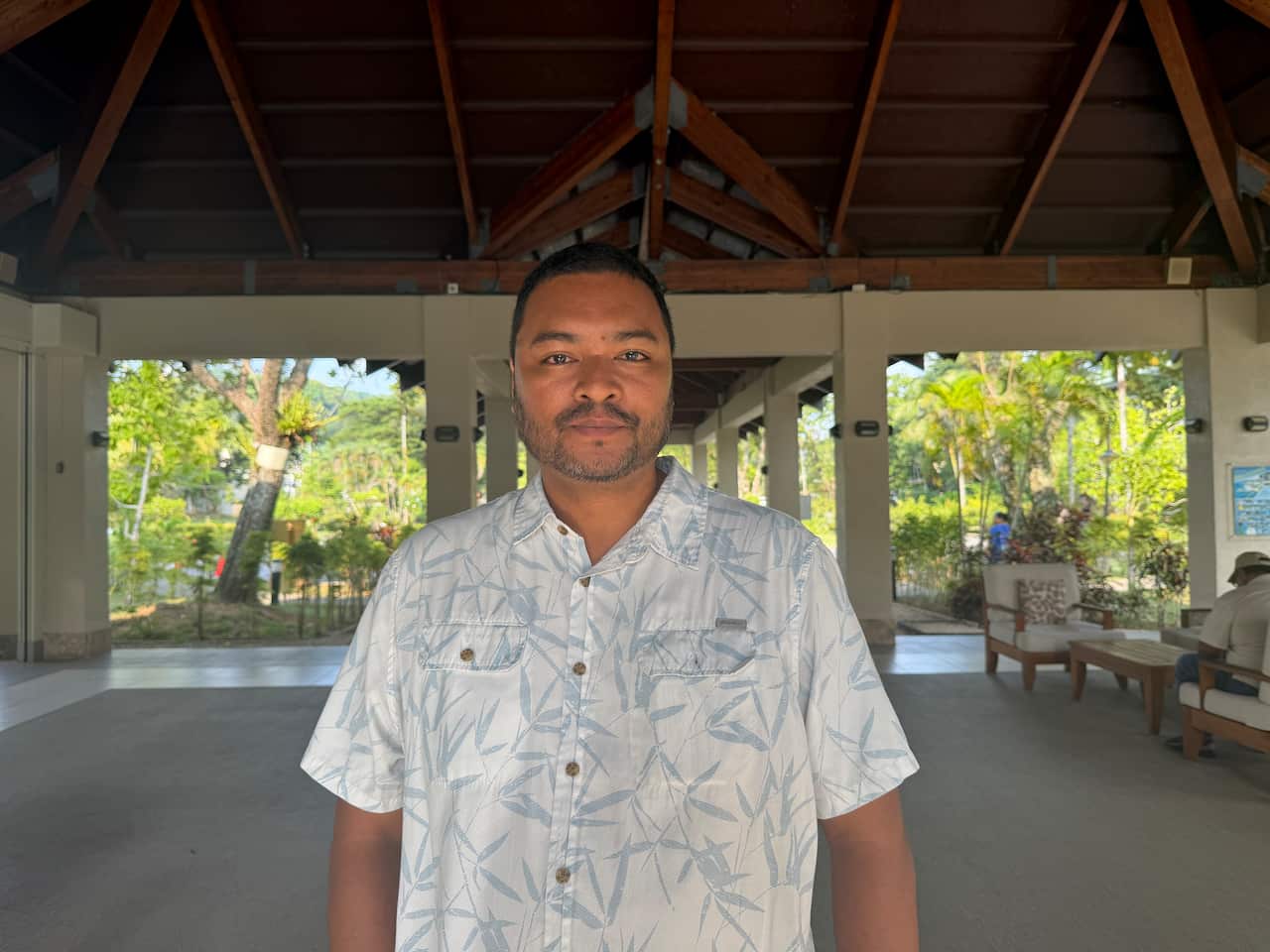 Man in a light patterned short-sleeve shirt stands inside an open-air resort lobby, with tropical gardens, seating and timber roof beams in the background.