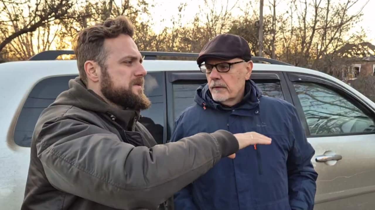 Two men standing outside a grey SUV. One is talking to the other and gesturing with his right arm as he does so.