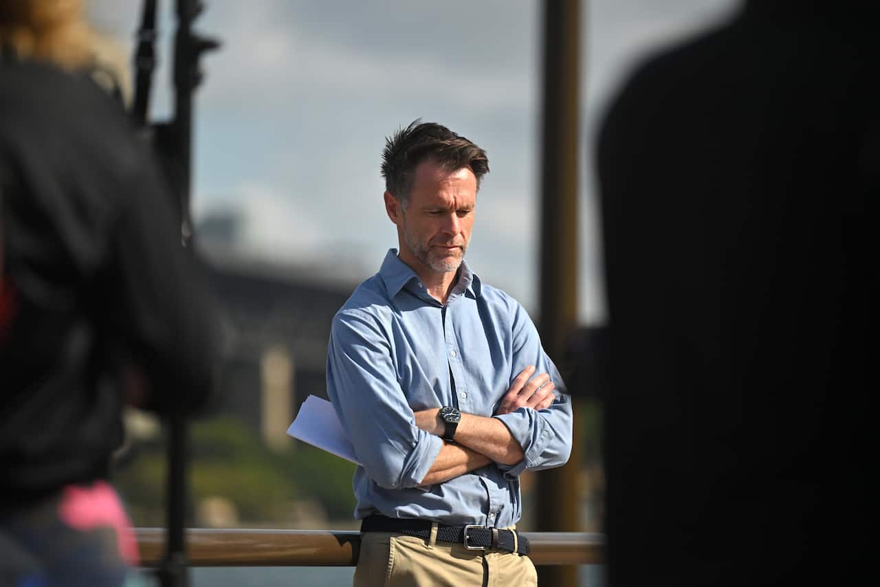 A man wearing tan chinos and a long-sleeved blue collared shirt stands with his arms folded.