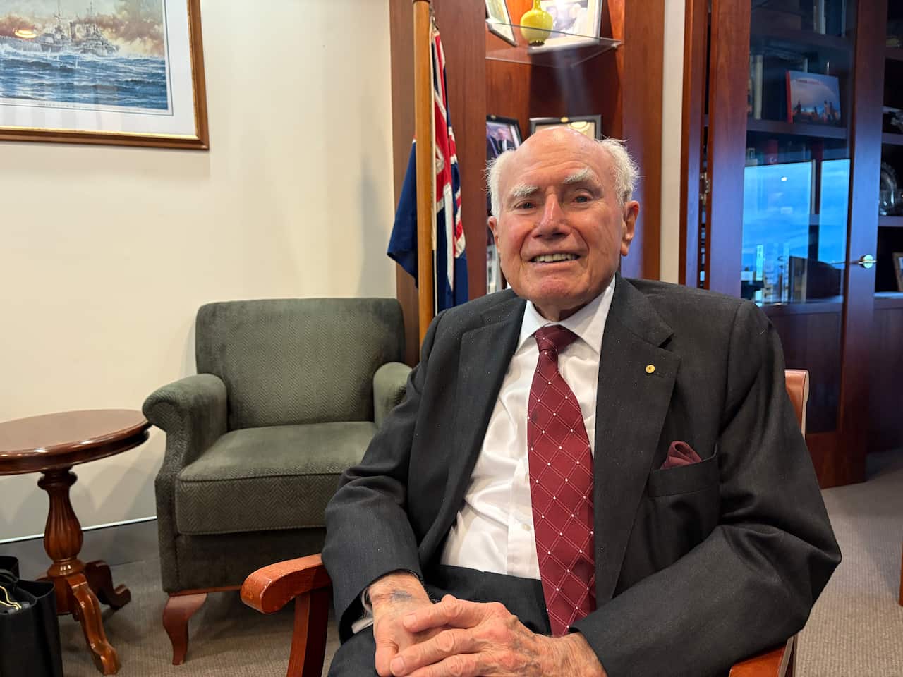 John Howard, in a dark suit and red tie, sits in a chair in an office.