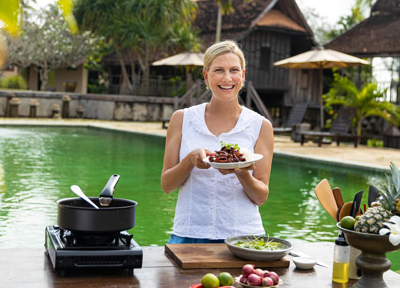 A similing woman in a white sleeveless top stands outdoors, in front of a pool, with a wooden building on the far side. She holds a bowl of food, and portable cooking stove and fresh ingredients sit on a table in front of her. 