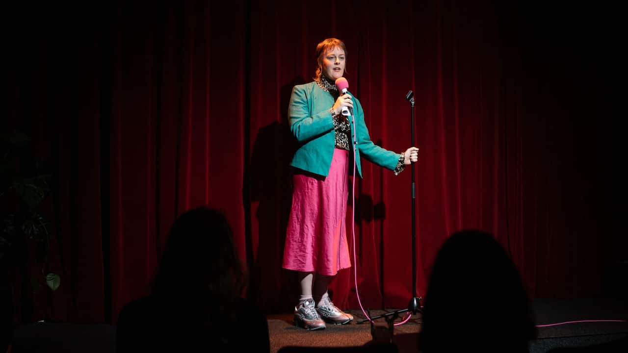 a young woman in a pink dress and blue blazer performs on stage holding a microphone
