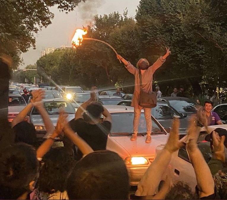 A woman stands on a vehicle burning her hijab. People applaud