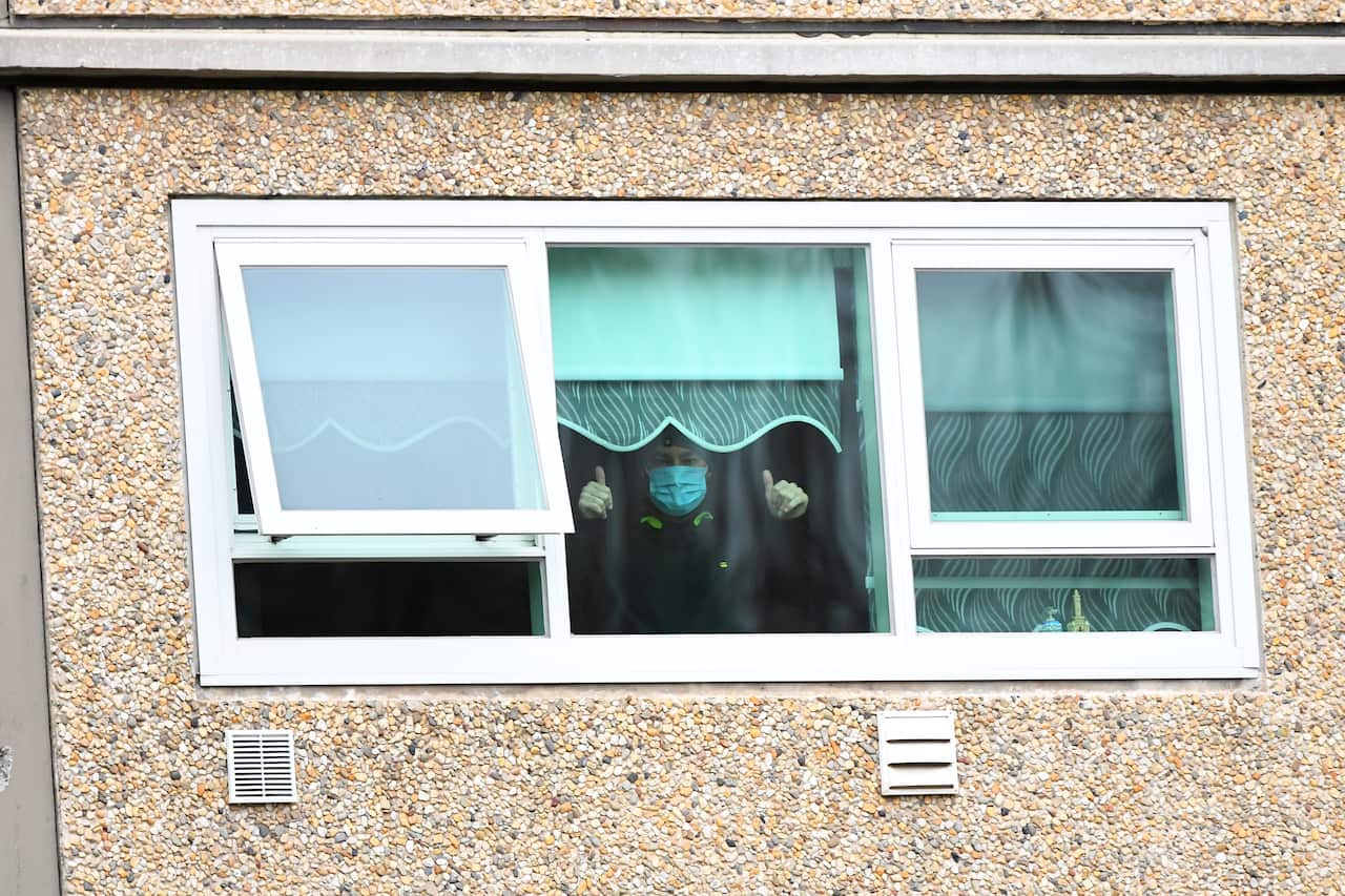 A man inside a window, wearing a mask holding both thumbs up.