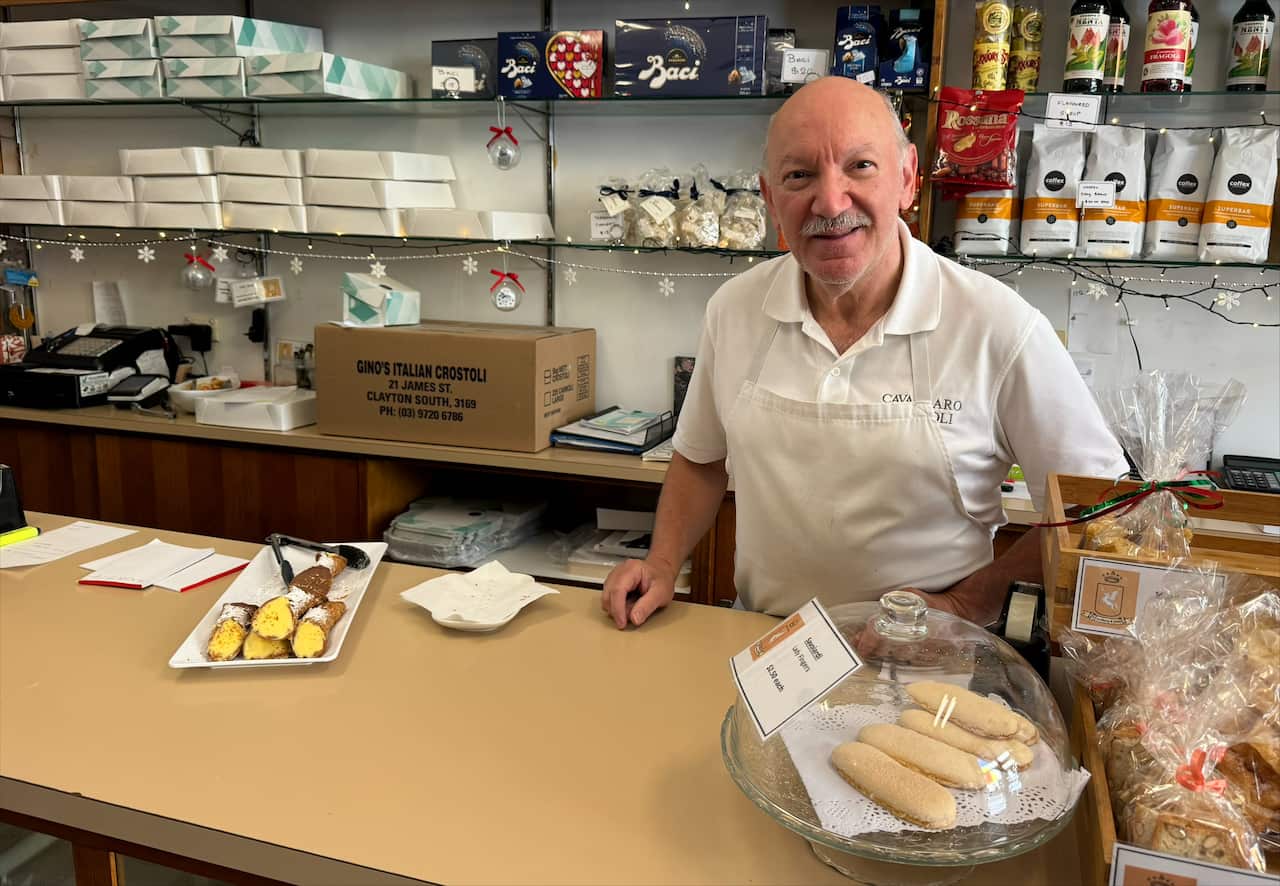 A man in a white apron stands behind a counter in a sweet shop, with shelves stocked with candy behind him.