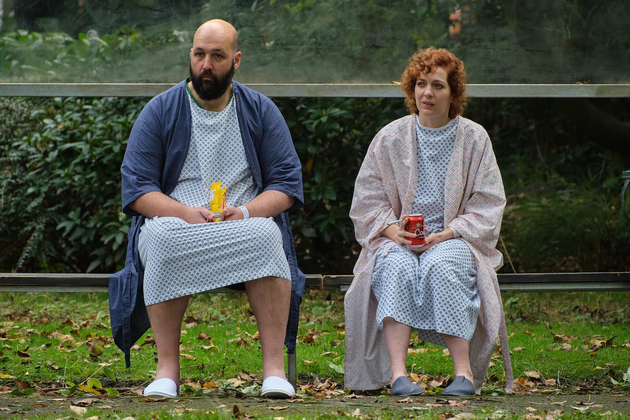 A man and a woman wearing hospital gowns sit next to each other in a garden