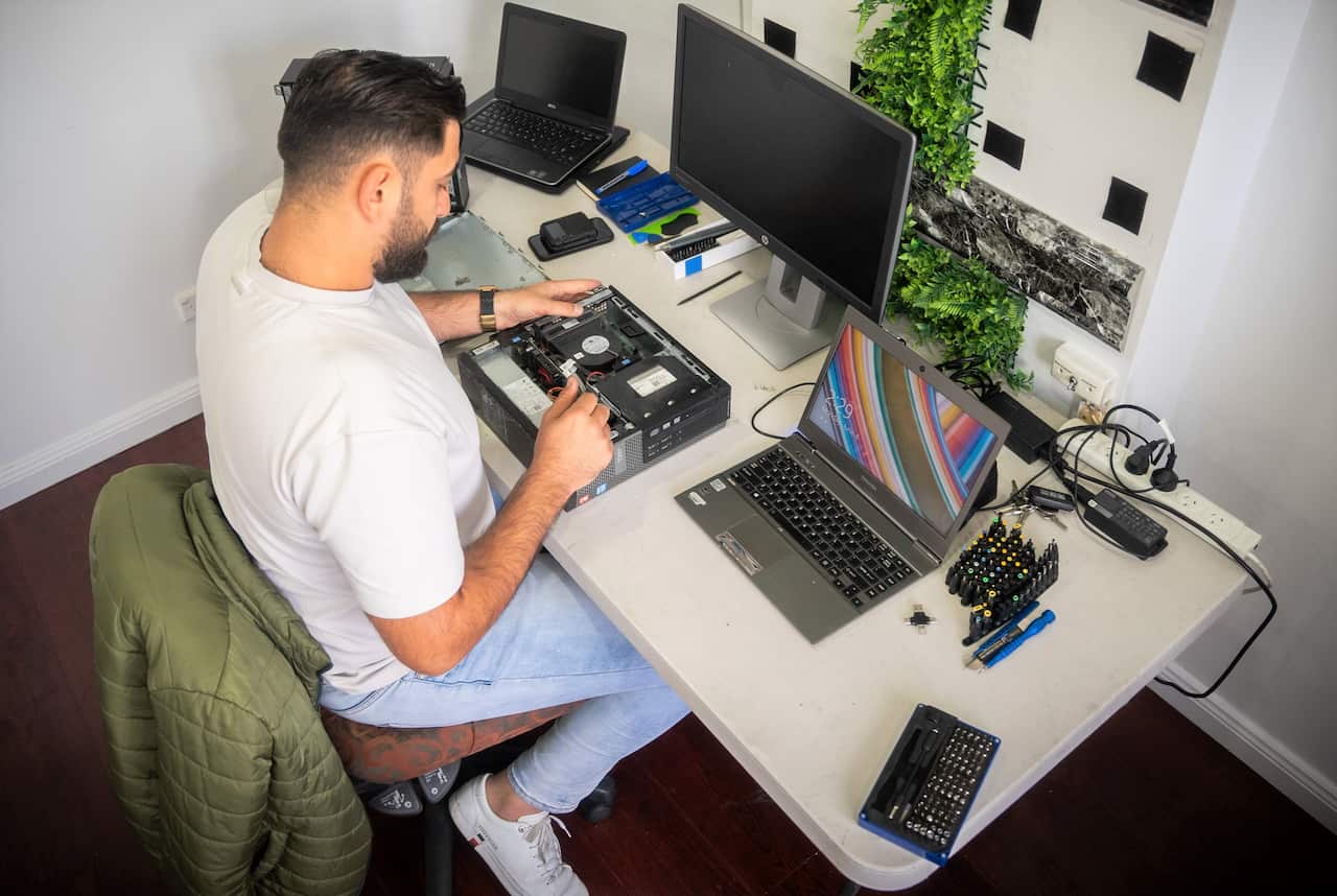 A man in a white t-shirt sits at a desk repairing a second hand computer.