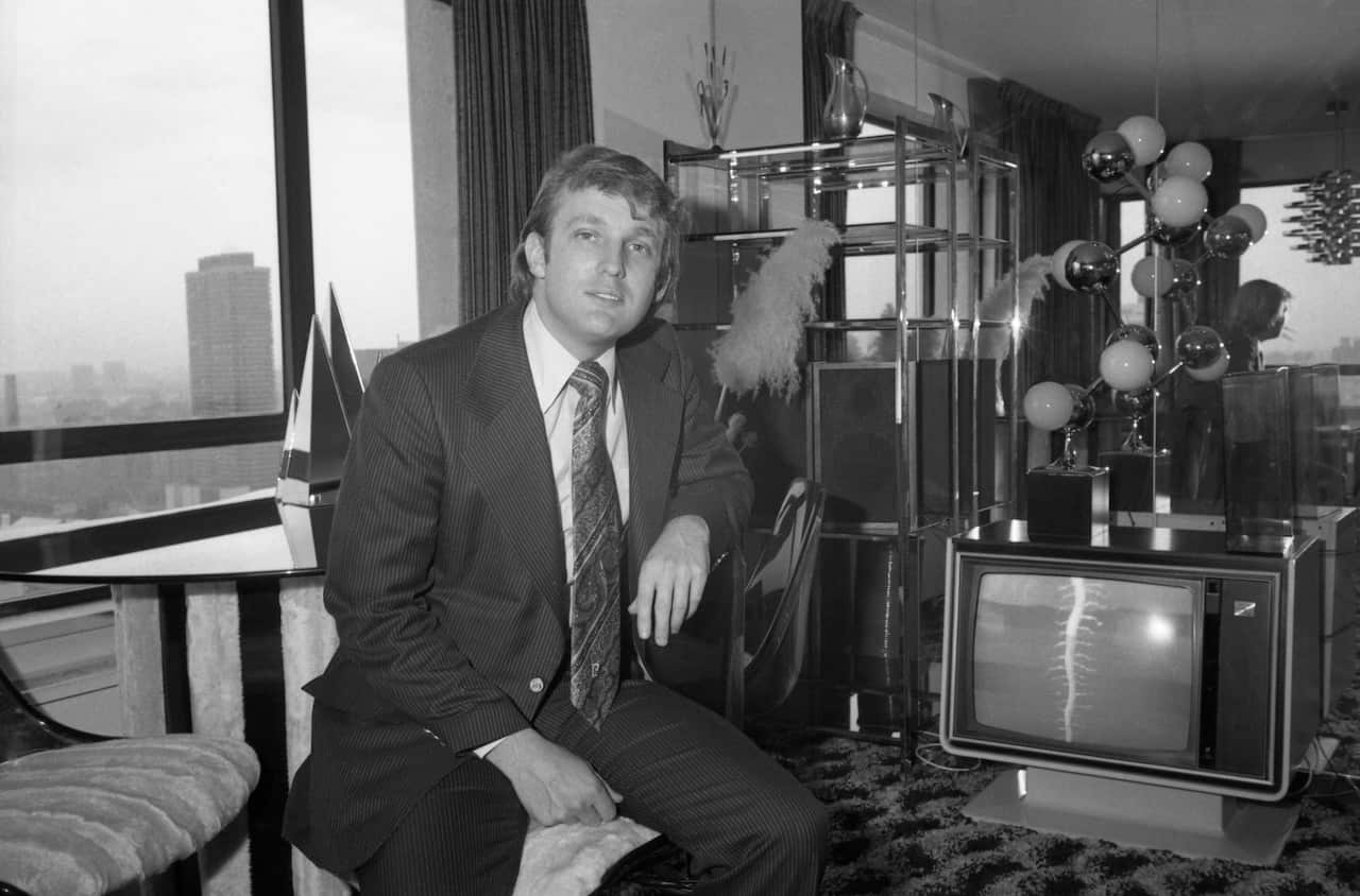 Young Donald Trump is seated in a room of a tall building, resting his arms on the back of a chair. A television stands beside him, and a shelf adorned with decorative vases is visible in the background.