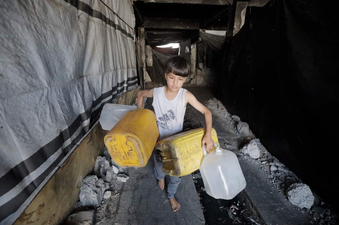 A barefoot boy in a white singlet carries four empty water containers through a rubble-filled corridor